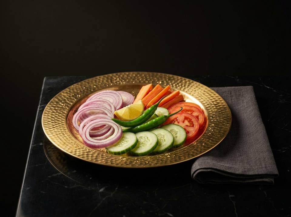 A brass plate of fresh salad: onion rings, cucumber and tomato slices, carrot sticks, green chillies and a wedge of lemon