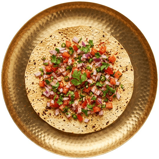 Seen from above: a masala papad topped with onion, tomato, green chilli and coriander on a brass plate